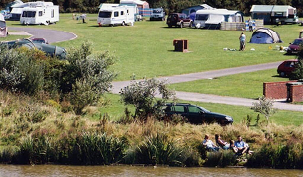 People camping on a grassy riverside park with caravans, tents, and parked cars