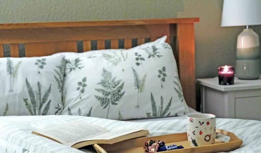 A bed with a tray holding an open book, a mug, and candy beside a lit candle on the nightstand