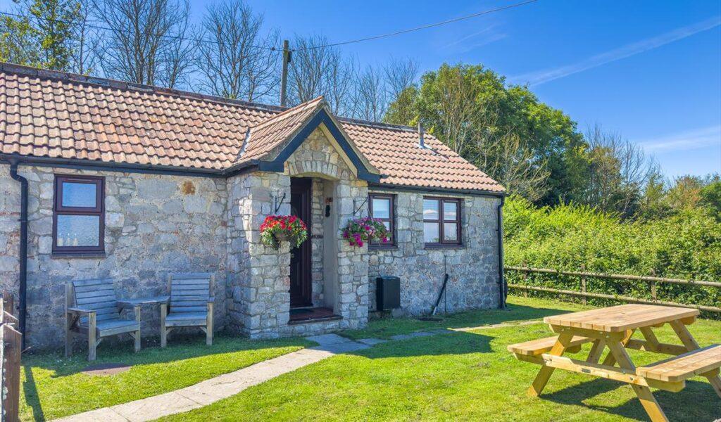 Stone cottage with a tiled roof, flower baskets, and a picnic table in a sunny garden
