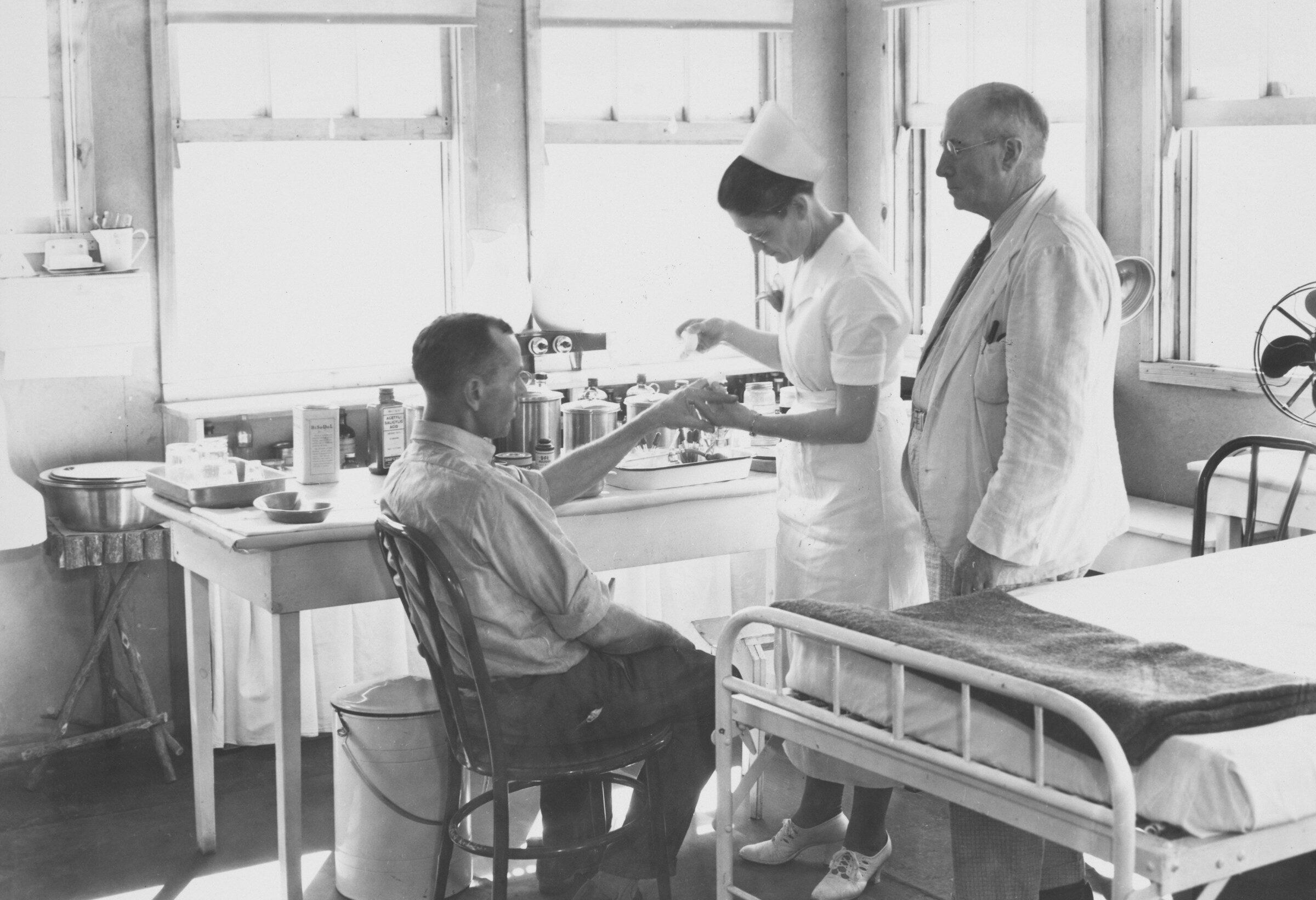 A black and white photo from the 1930s of a doctor and nurse attending to a man sat in a chair
