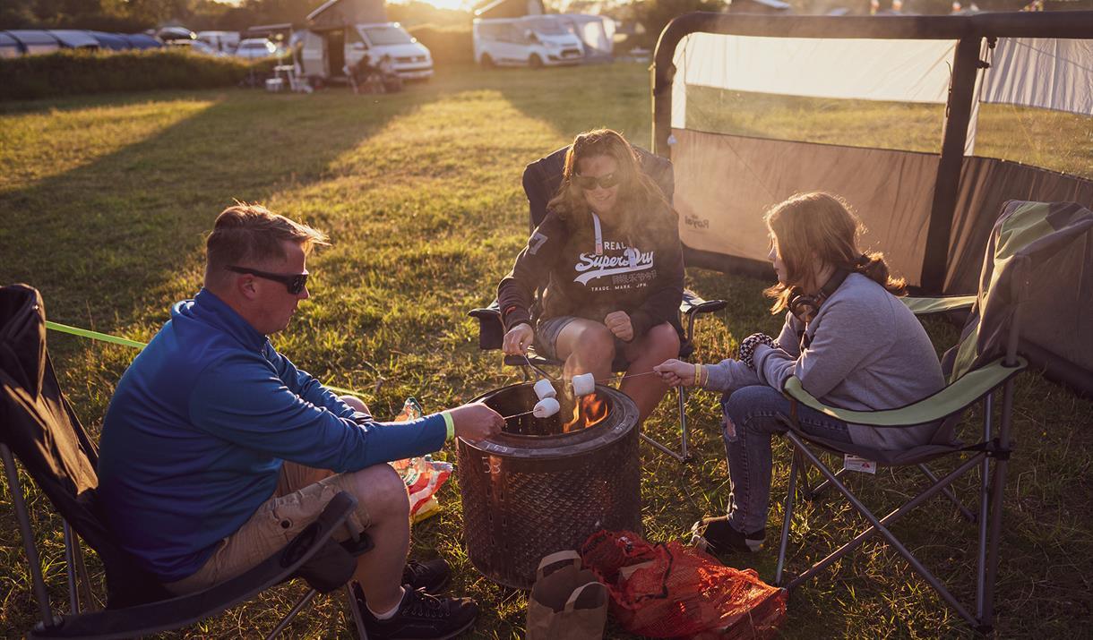 A man, woman and girl sit in camping chairs around a controlled fire in a metal drum, toasting marshmallows on sticks