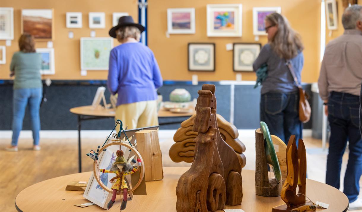 A collection of wooden and fabric art pieces sit on a table. Behind that 4 people study the walls and a second table, which feature multiple other art works