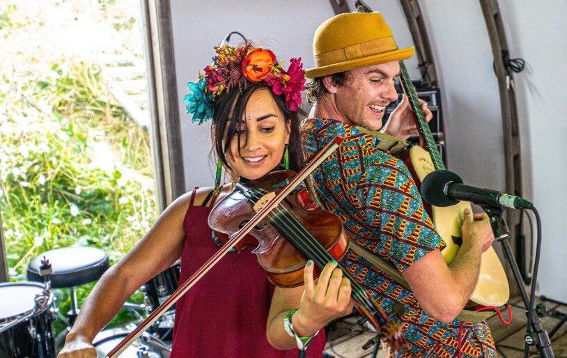 A woman with flowers in her hair plays the violin. A man with a yellow bowler hat plays a guitar. Behind them is the inside of a tent.