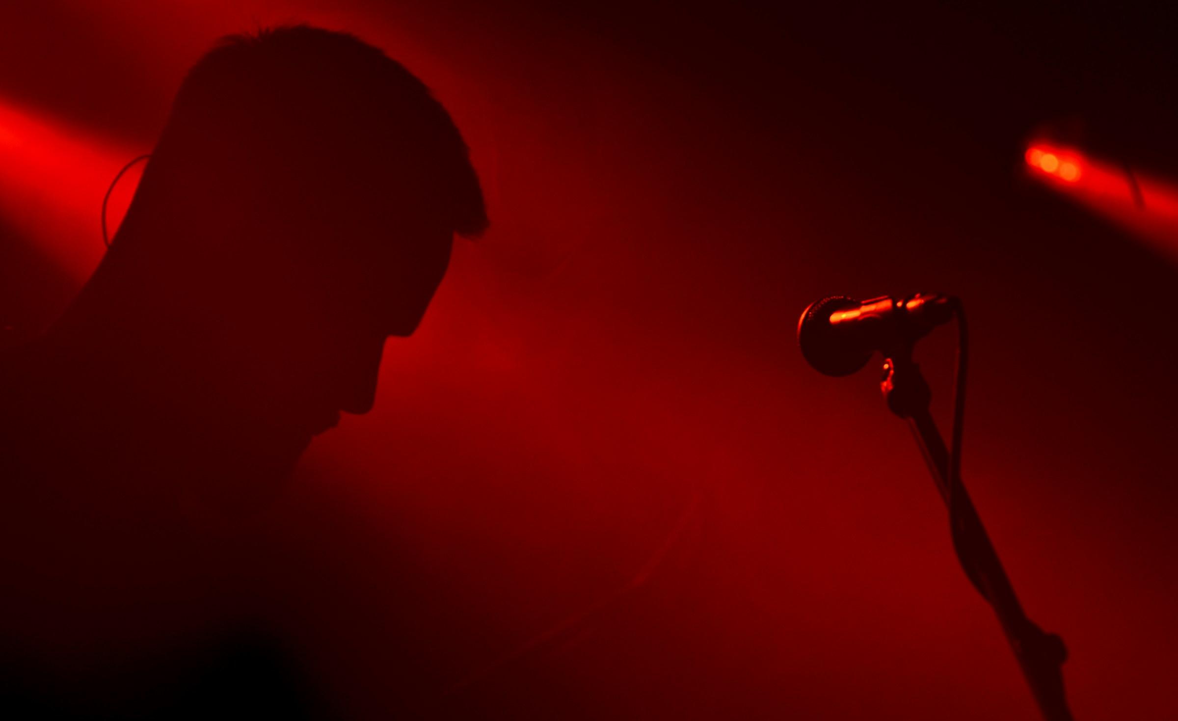 Over a red and black background stands a microphone stand and a man in silhouette.