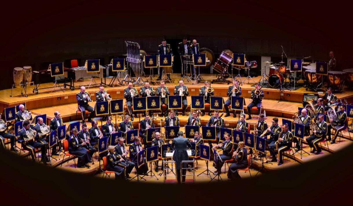 A large group of musicians all wearing blue uniforms sit in a half semi circle around the conductor.