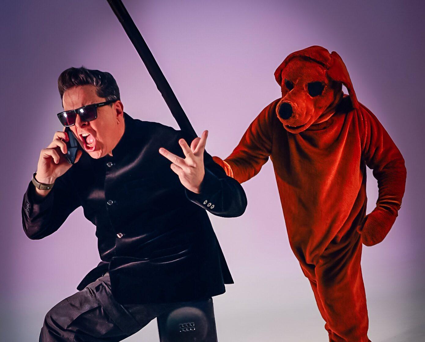 A man in dark clothing and sunglasses holds a small phone to his cheek, while he sits on a large phone. A person in a red dog costume is mid way through hitting the man with a stick.