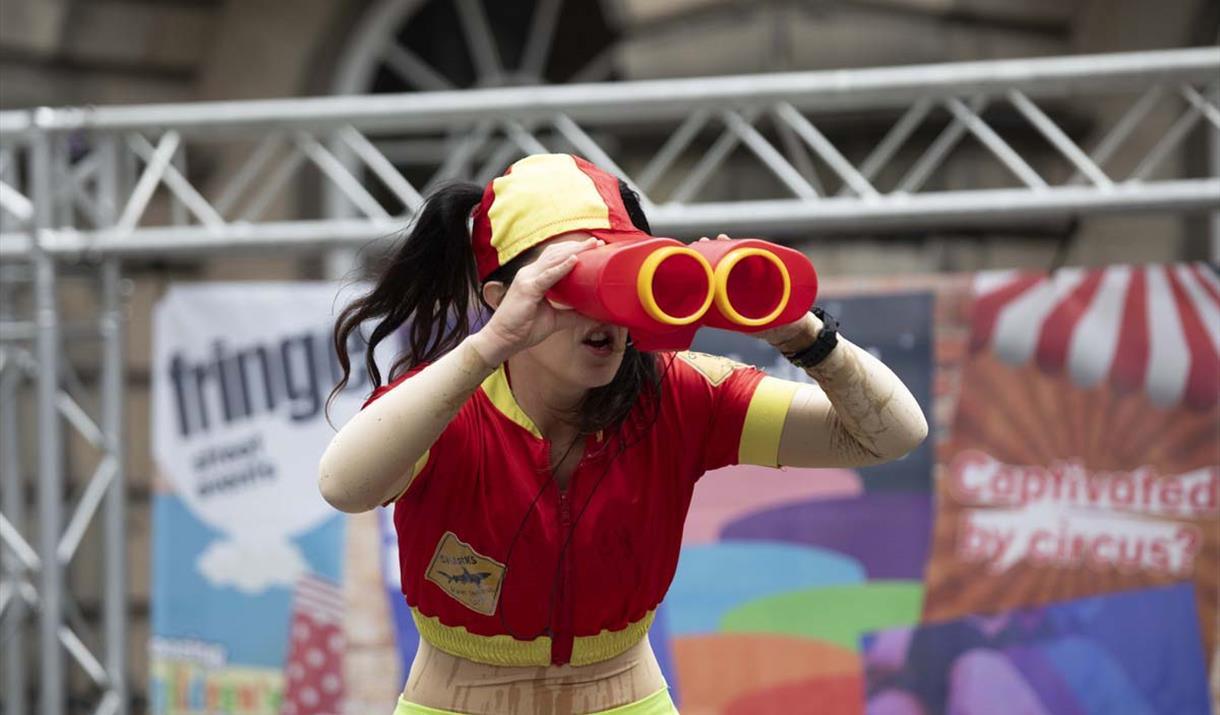 A performer at Whirligig. A woman with a bright red and yellow outfit holds red and yellow binoculars in front of her face.