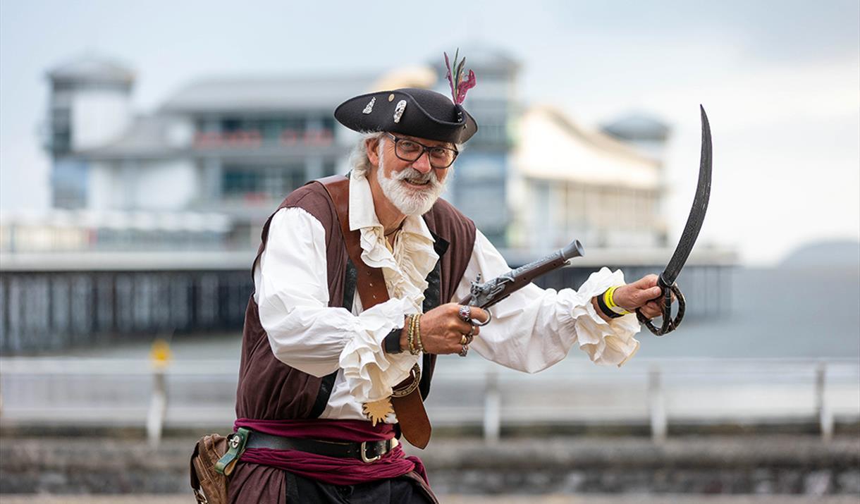 A man stands in a full pirate outfit with a fake cutlass and gun. Behind is the Grand Pier
