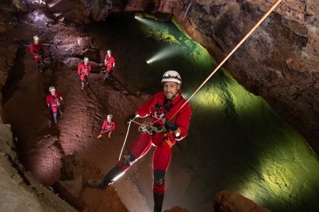 A man in a red boiler suit abseils into a cave to join more climbers o the Wild Wookey Experience