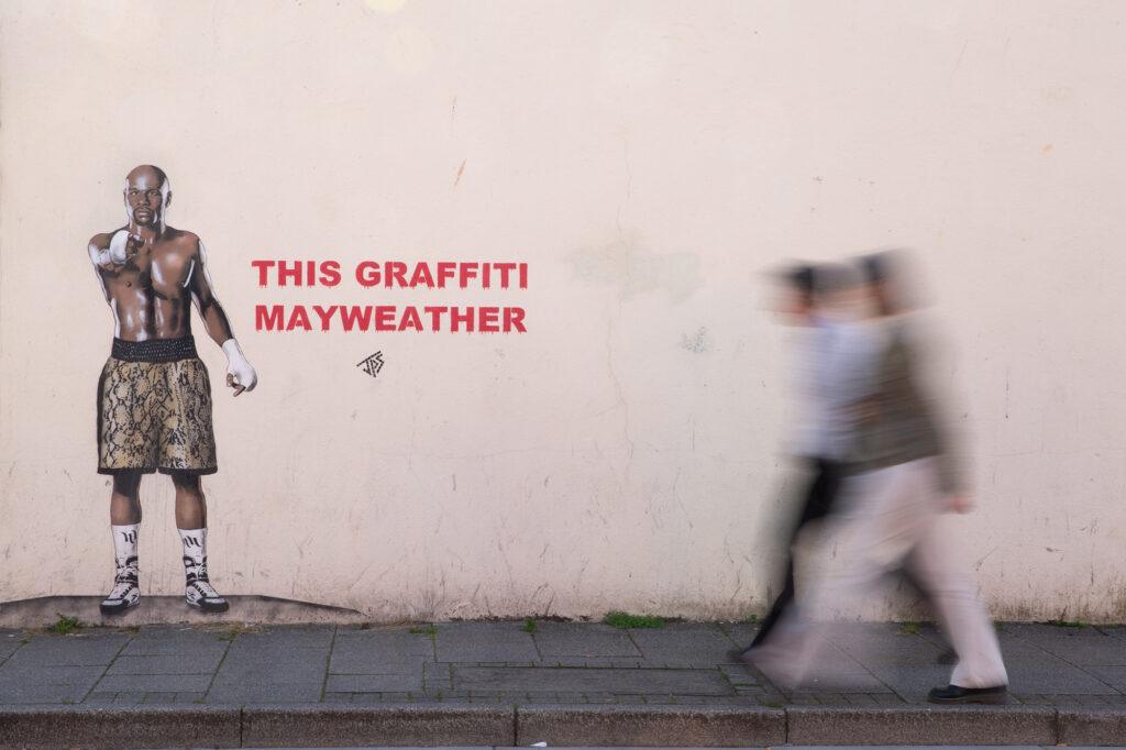 Two walkers whose motion is blurred stroll past a mural featuring boxer Floyd Mayweather in Weston-super-Mare with the words This Graffiti Mayweather