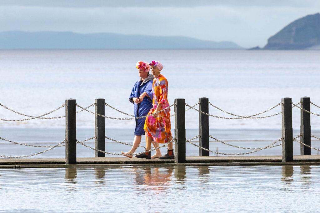 Two swimmers in swimming hats and robes walk along the causeway at Weston-super-Mare's Marine Lake