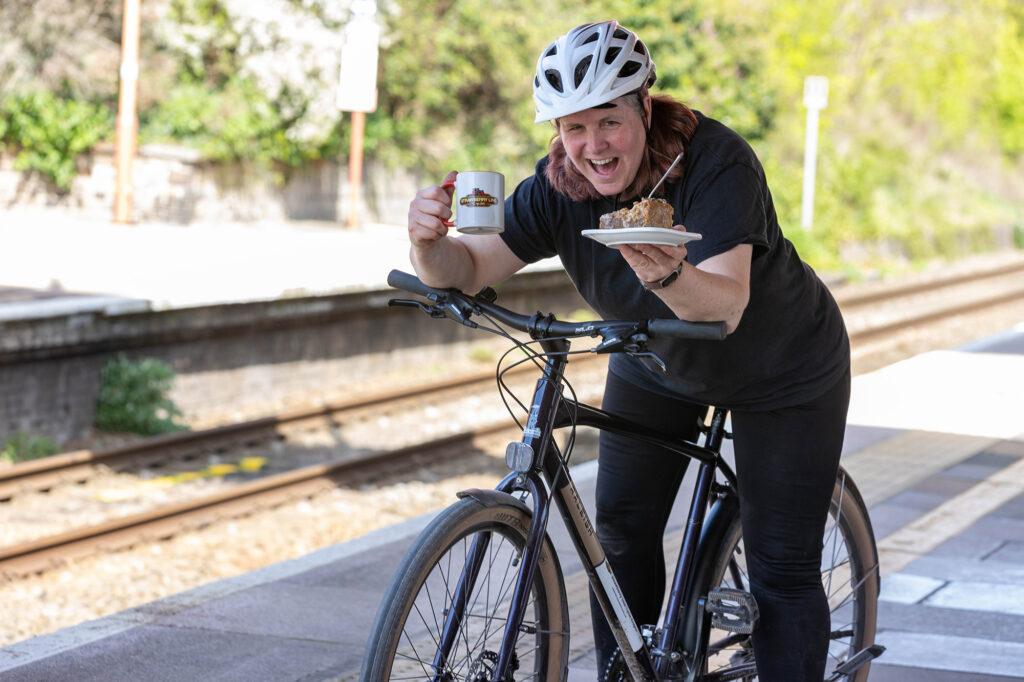 A cyclist sitting on her bike on Yatton railway station eating a slice of cake and drinking a mug of team from the station's Strawberry Line Cafe