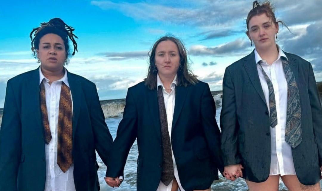 3 women dressed only in a shirt, blazer and tie each stand on a beach in front of the sea