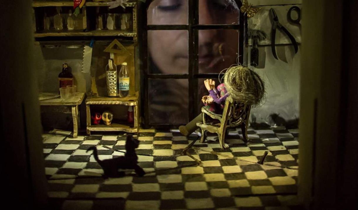 A real life person looks in through the window of a small puppet kitchen, with a cat and a woman on a rocking chair inside.
