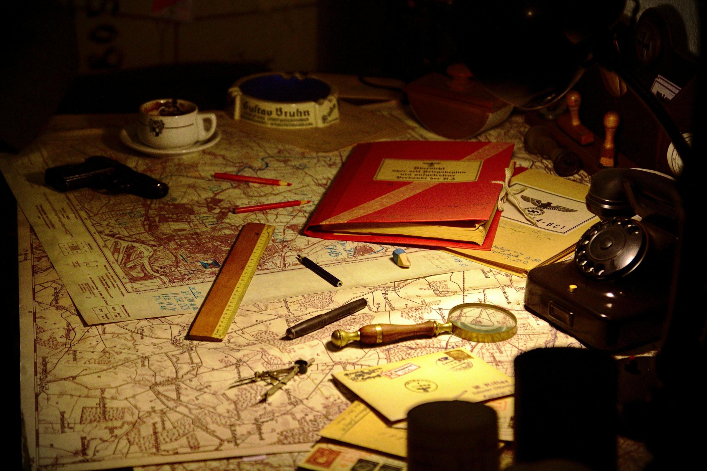 On a table sits a map, rulers, pencils, documents, a coffee cup and a gun