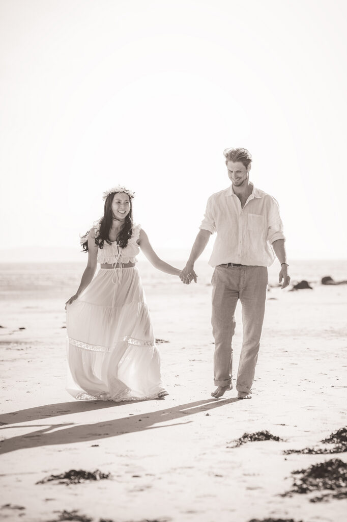 A high key black and white image of a man and woman strolling hand in hand on a beach