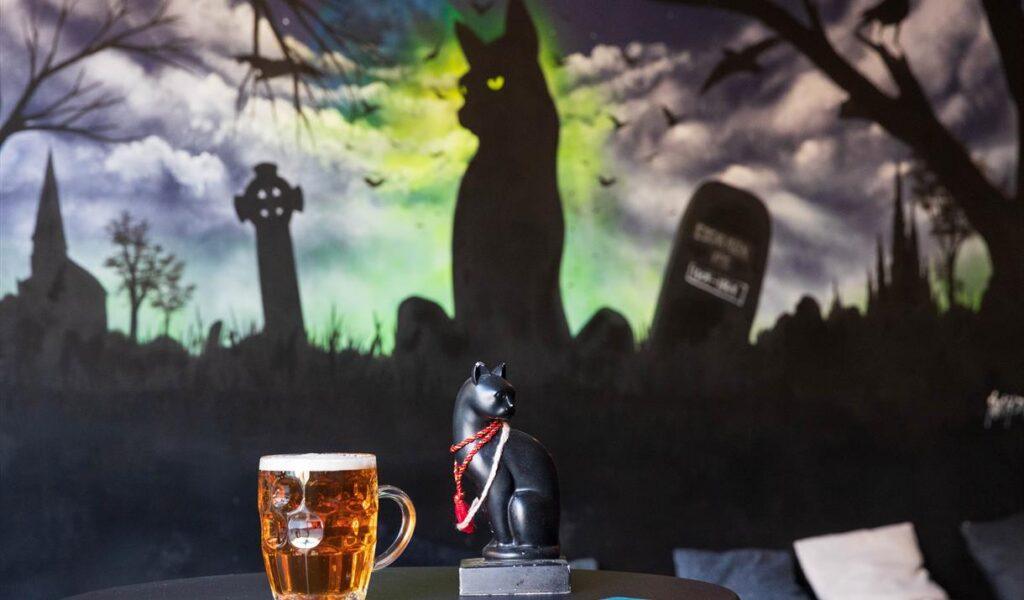 A Pint of beer and a black cat figurine on a table in front of a mural of a black cat in a graveyard