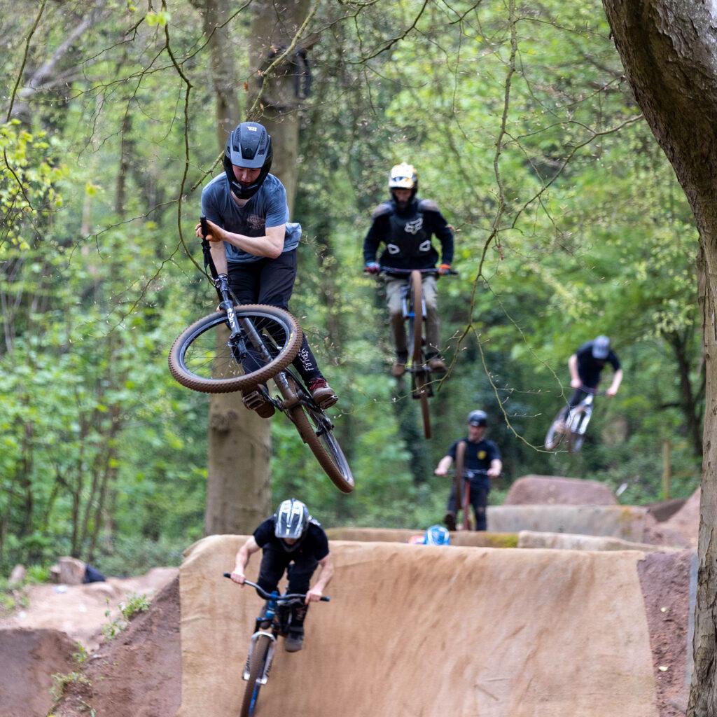 Five cyclo-cross riders, three of which are airborne, taking on the Sand Bay Dirt Jumps in Weston Woods, Weston-super-Mare.
