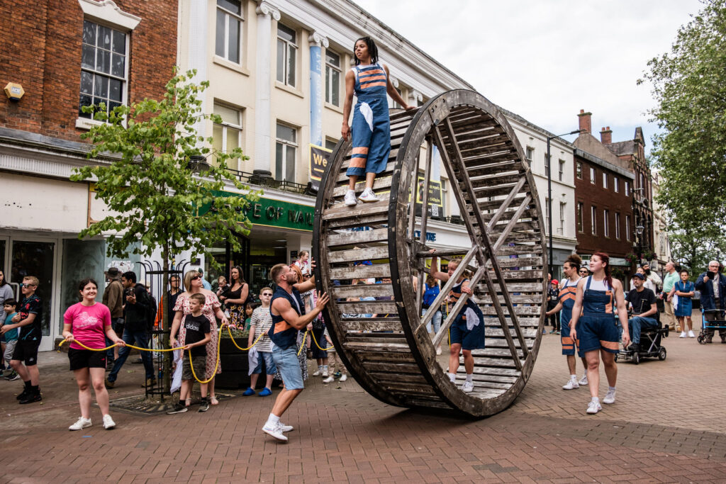 A woman in blue dungarees walks on a giant mill wheel which is being rolled down a busy shopping street by her colleagues who are supporting the wheel.