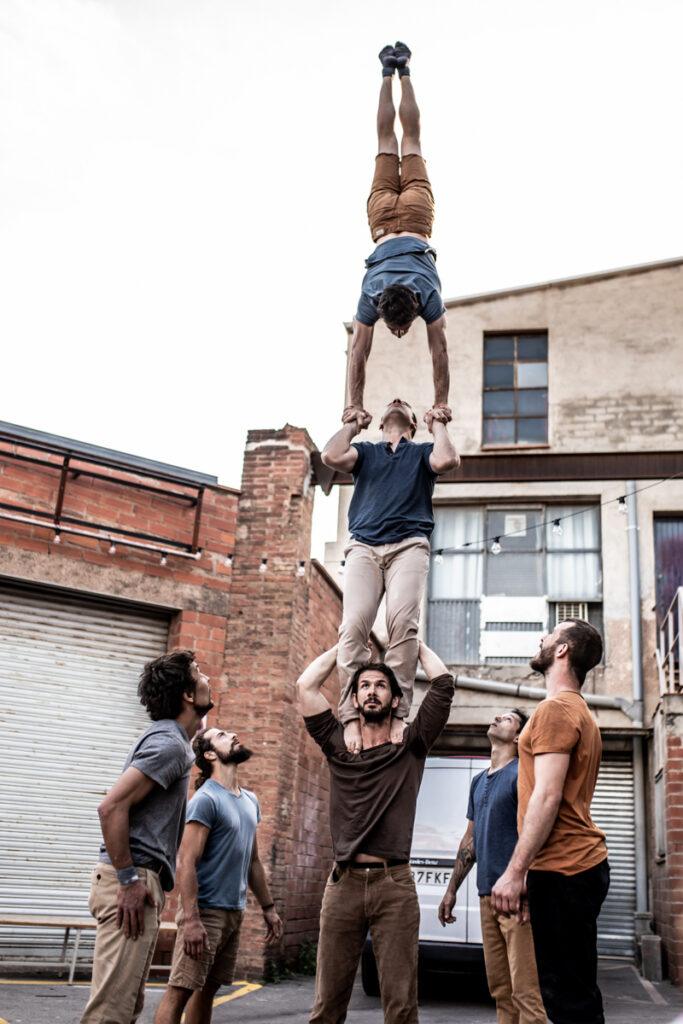 A human tower of three men reaching high into the sky as four other men look upwards at this unusual creation near two lock up garages