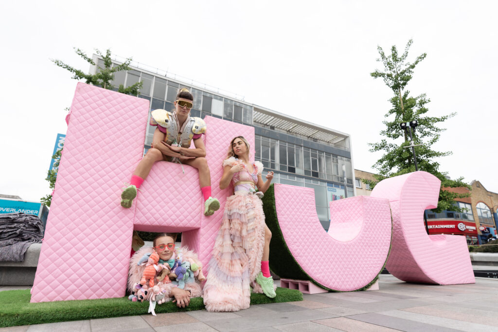 Three giant pink letters spelling Hug with two women in flamboyant costumes sitting on and under the H while a thrid performer leans on the letter H