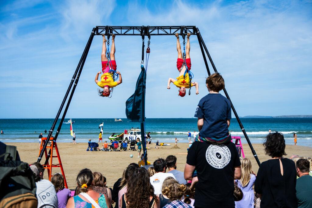 A crowd watch a performance on a beach of two lifeguards hanging down from what looks like a giant swing