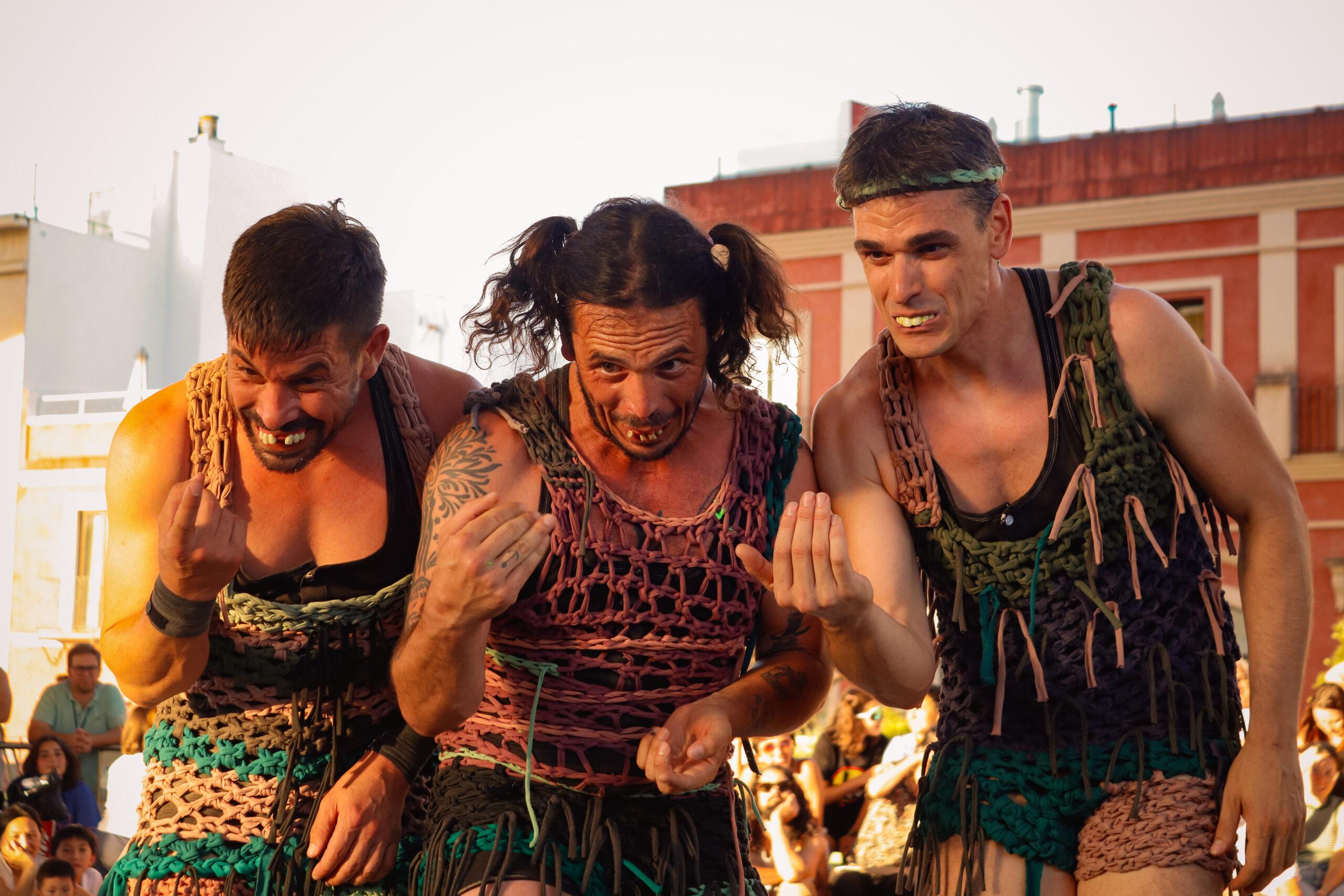Three performers in string vests and with their teeth protruding hold out their right hand and look downwards