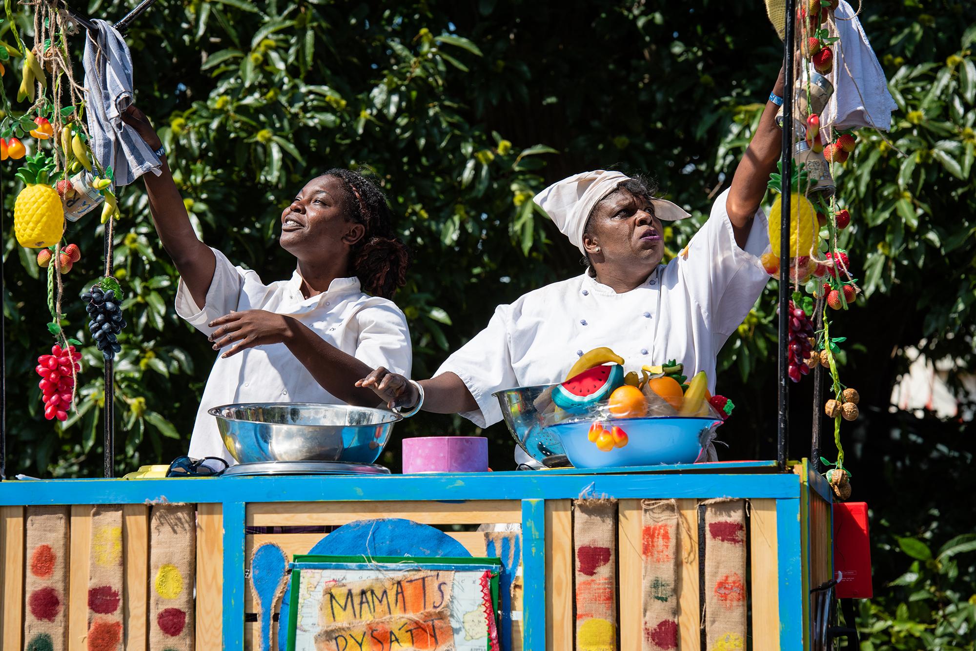 Two women standing behind a food stall and wearing chef's whites whites reach upwards
