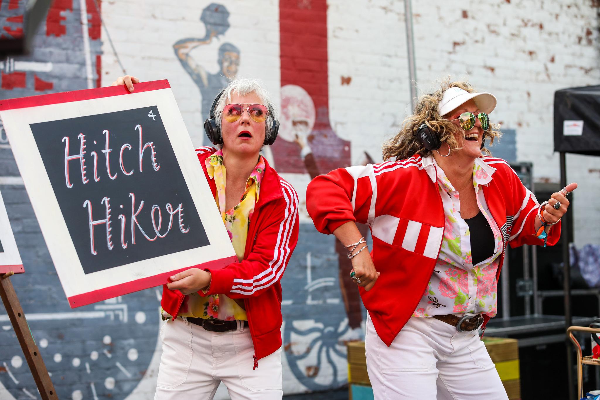 Two women wearing red jackets and white trousers are dancing with their headphones on while holding up a board saying Hitch Hiker