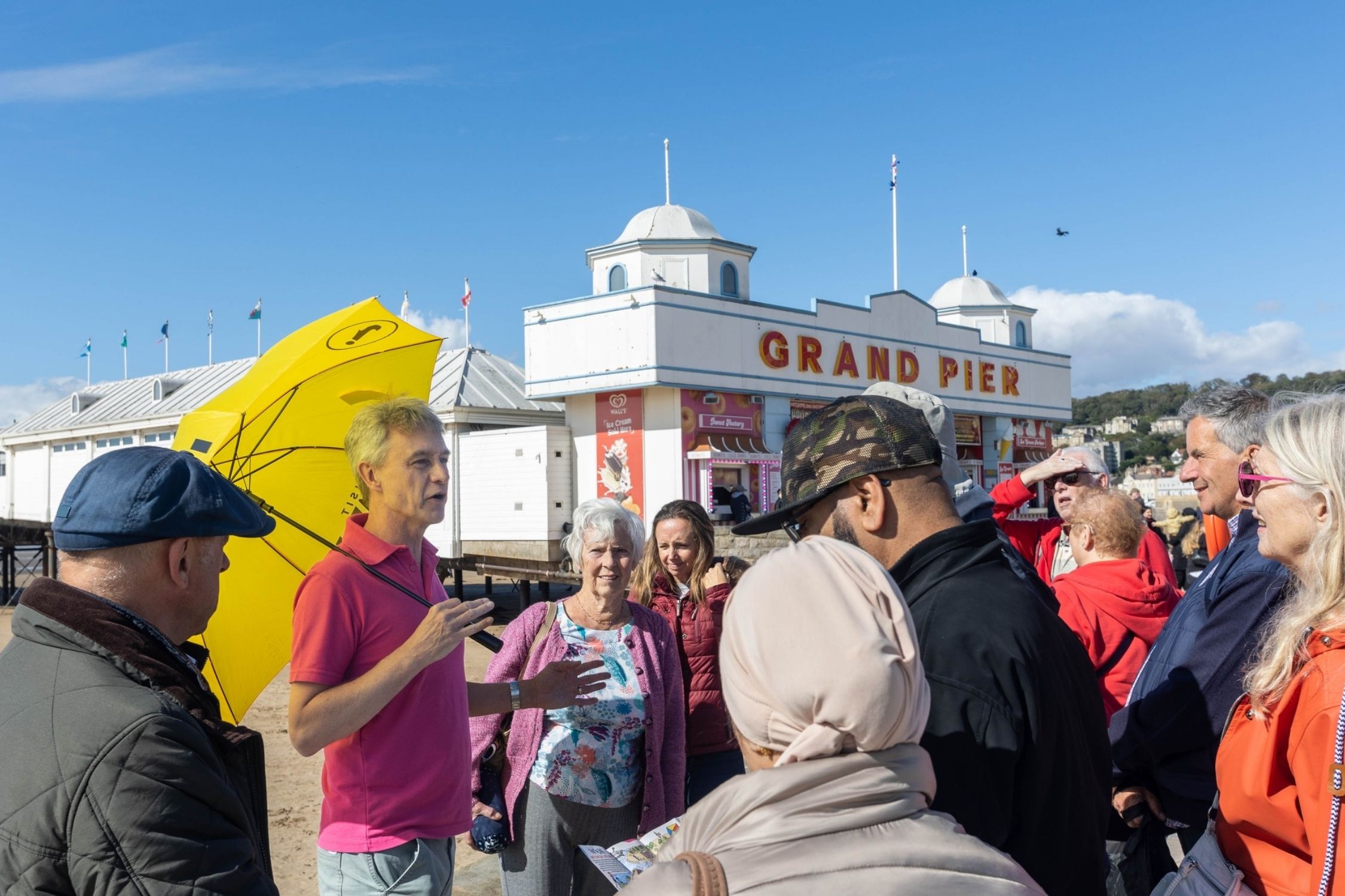 A man in a pink T-shirt and yellow umbrella addresses a crowd of people on a walking tour outside the Grand Pier Weston-super-Mare