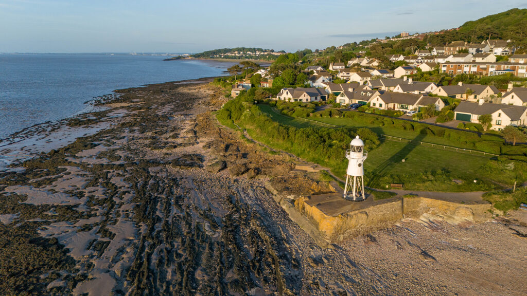 Aerial view of the white-coloured Black Nore Lighthouse and the rocky beach it sits next to with town of Portishead in the background