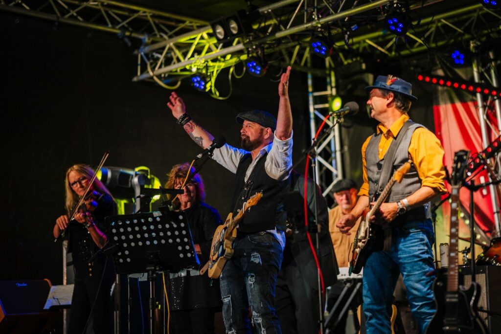 The lead singer of a four-piece folk band raises both arms out wide in appreciation of the cheers of the audience during a nighttime concert