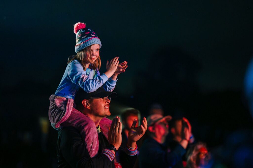 A girl, sitting on an adult's shoulders, claps as she applauds the show at a nighttime concert