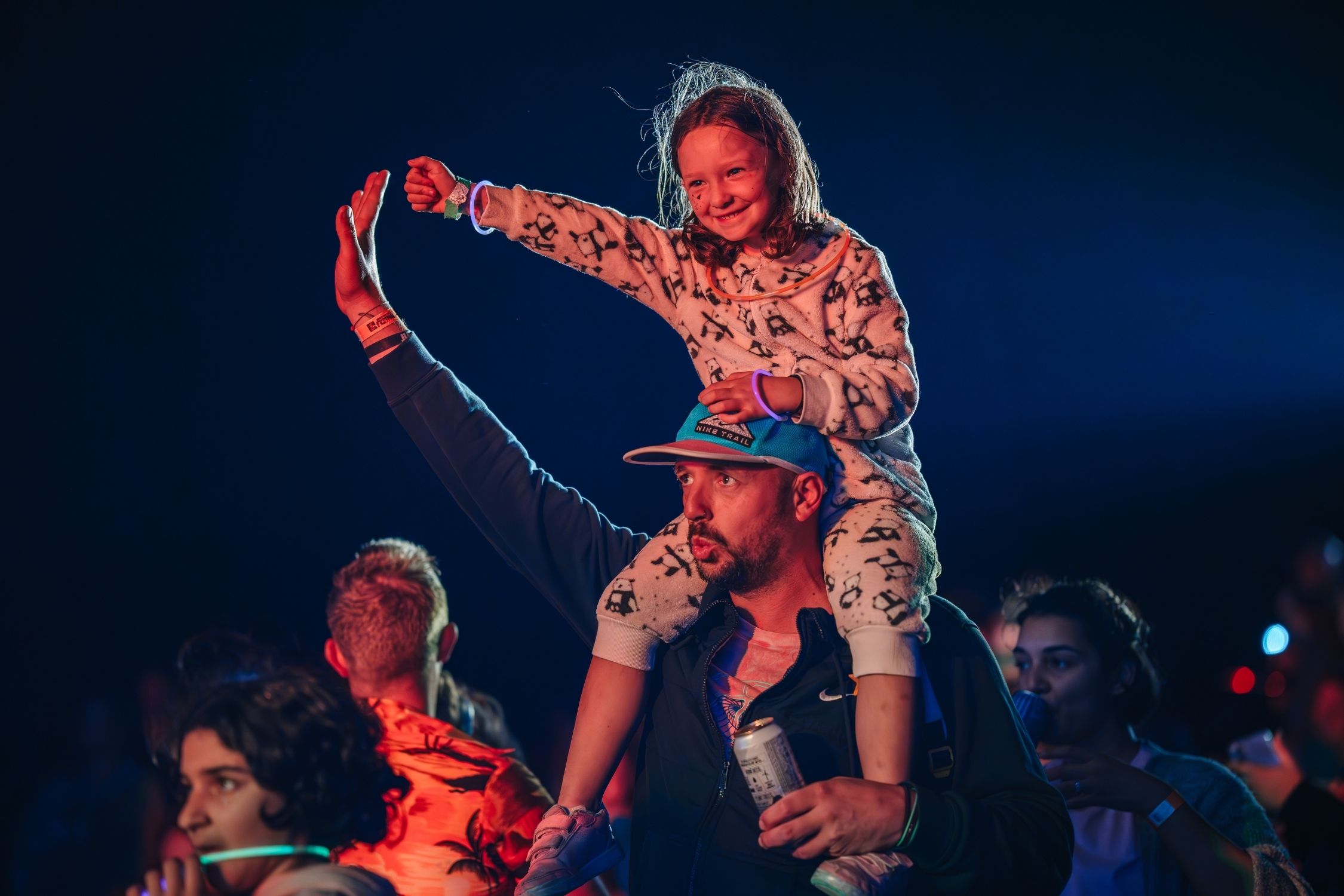 A dad has his young daughter on his shoulders as they both wave their arms while watching a nightime concert during a music festival