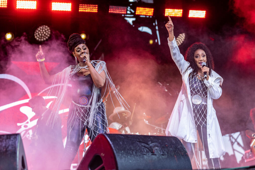 Two members of a girl band point their fingers in the air while on stage