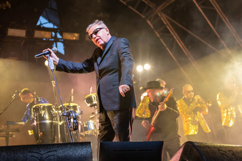 A lead singer in dark suit and sunglasses leans the microphone forward while his gold-jacketed band perform behind him