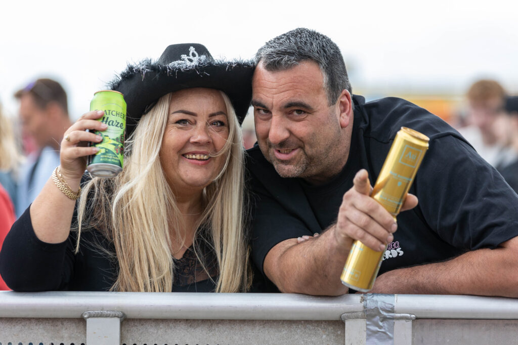 A man and woman dressed in black and holding drink can pose for the camera while leaning on a fence