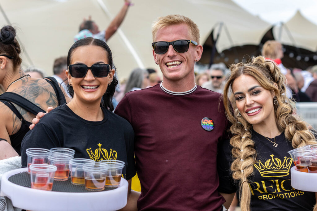A man in sunglasses in between two ladies holding trays of drinks