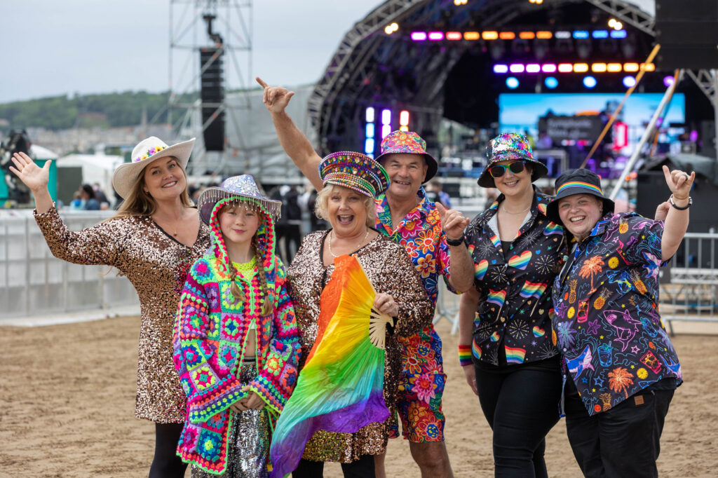 Six people in very bright clothes pose in front of a festival stage