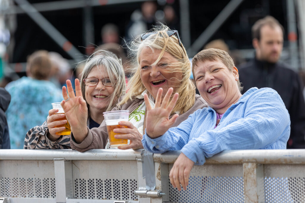Three ladies, two with drinks in their hand leaning on a fence and waving