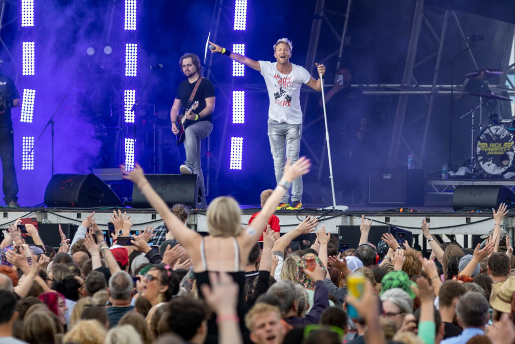A lead singer points to a large crowd at a festival which includes a girl on someone's shoulders with her arms outstretched