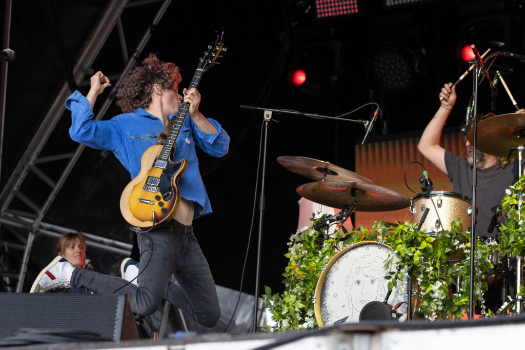 A male guitarist in a blue shirt and black trousers jumps and lands on his knees