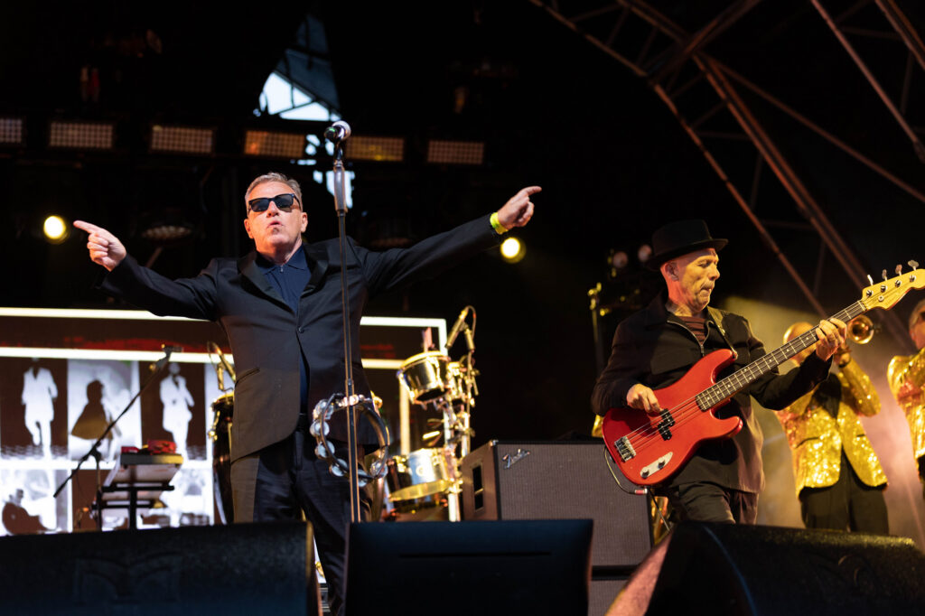 A dark suited lead singer with sunglasses on with his arms outstretched