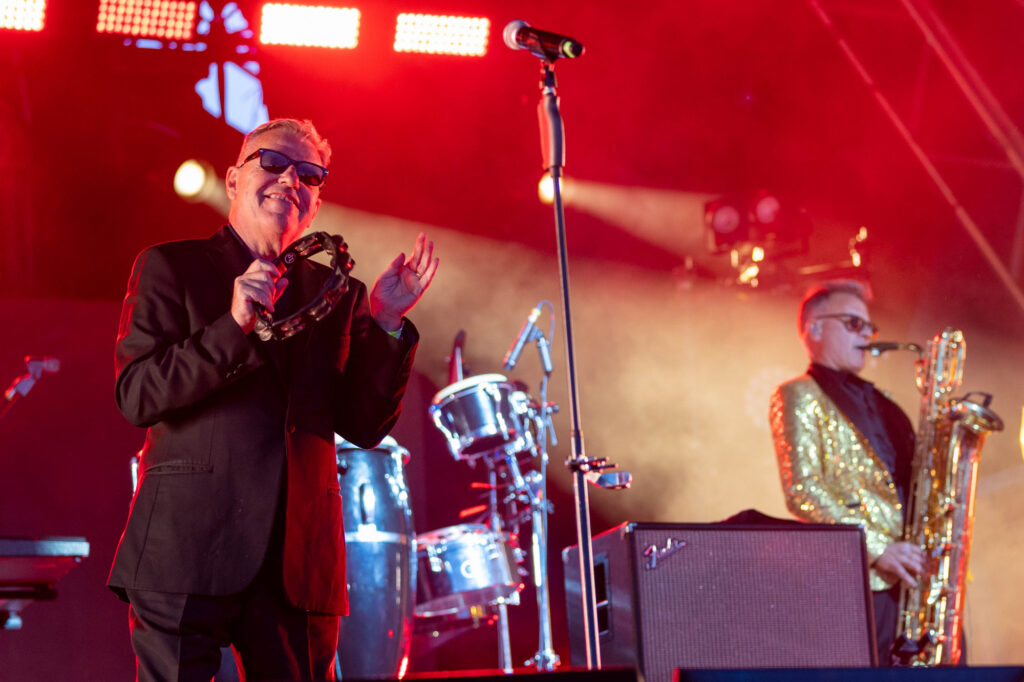 A man in a black sunglasses and suit plays tambourine on stage