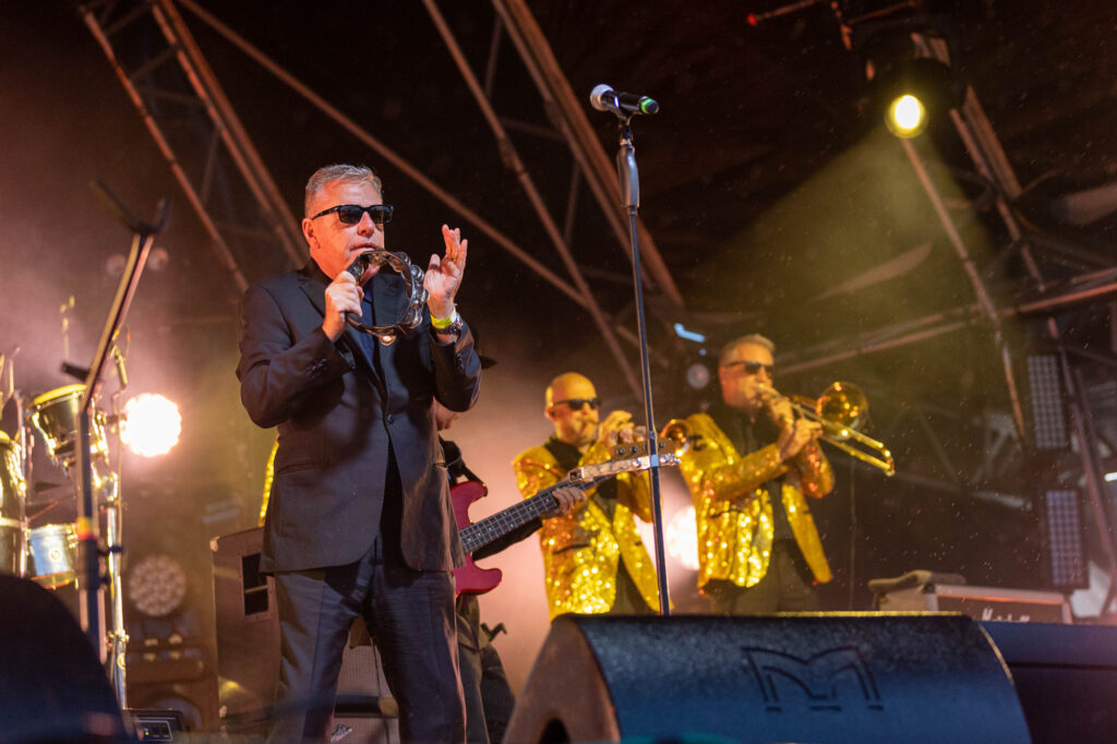 A singer in black suit and sunglasses plays the tambourine while his gold-jacketed colleagues perform in the background