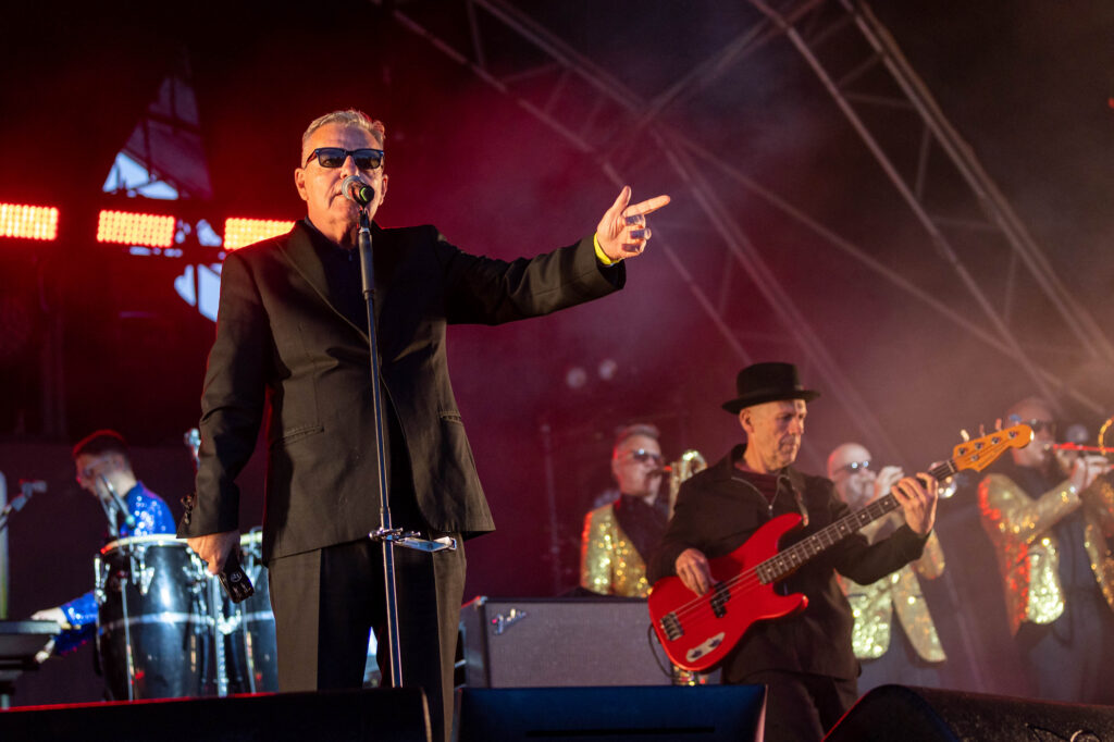 A singer in sunglasses and a black suit points to his fellow band members