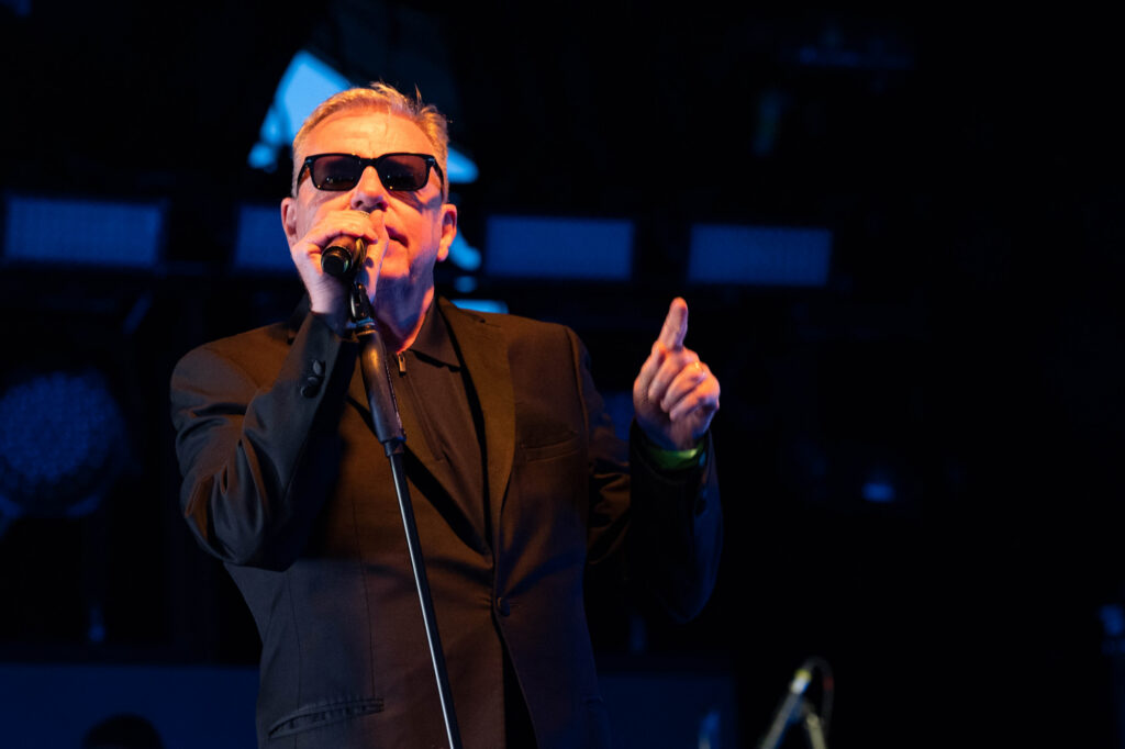 Suggs of Madness wearing sunglasses and a black suit holding a microphone and pointing