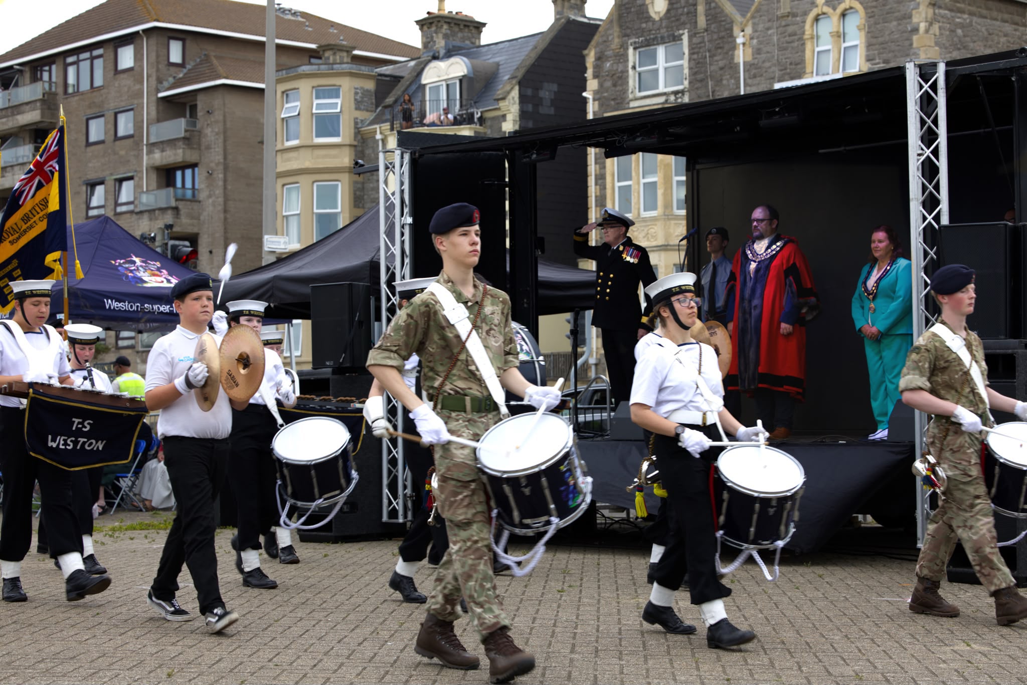 Drummers in army cadet and Sea Cadet uniform march past a tented stand housing a major and a senior military officer