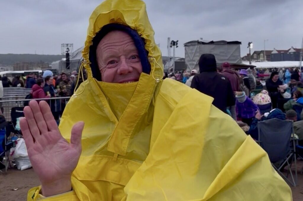 A man at a music festival in a huge yellow coat gives a cheery wave as the rain comes down