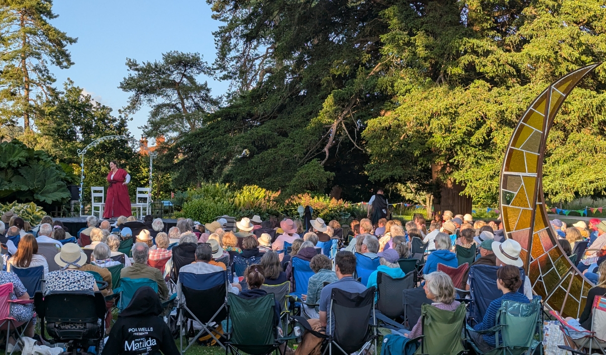 A crowd of people sit in a large garden space, watching an actor perform